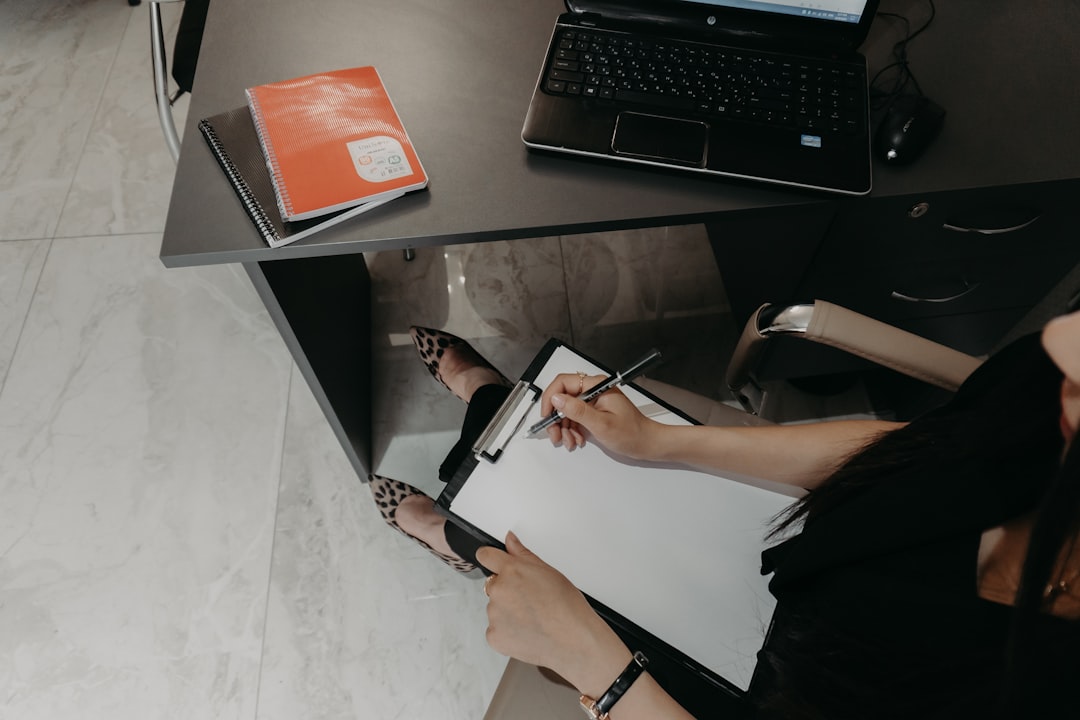 A person marking up a document on a clipboard beside a laptop at a desk